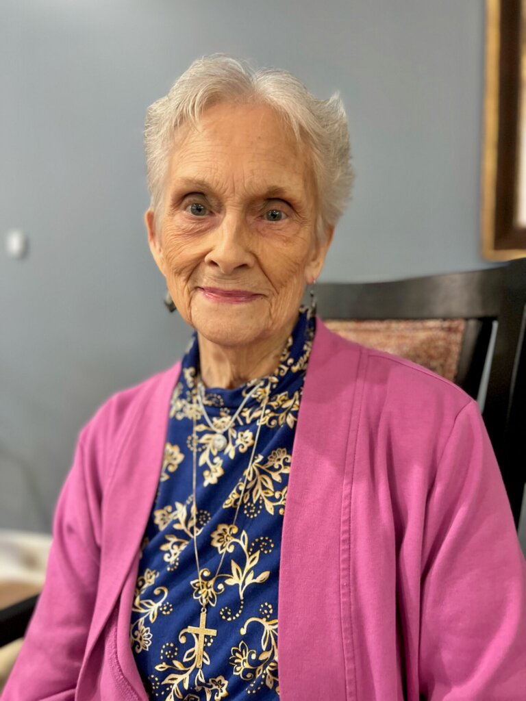 A close-up of an older woman wearing a pink cardigan and blue patterned top.