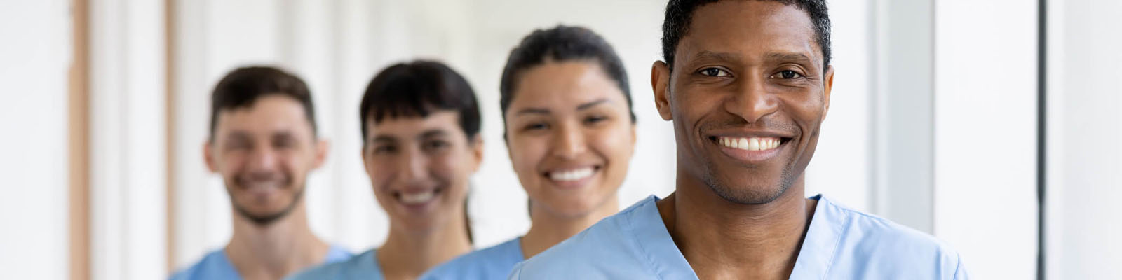 Four senior living nursing assistants wearing blue scrubs staggered behind each other in a line smiling
