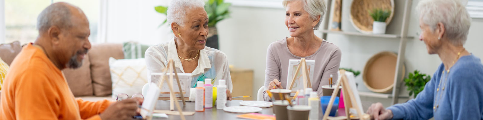 Male and female senior living residents sitting at a wooden table with mini easels holding small paintbrushes and painting pictures