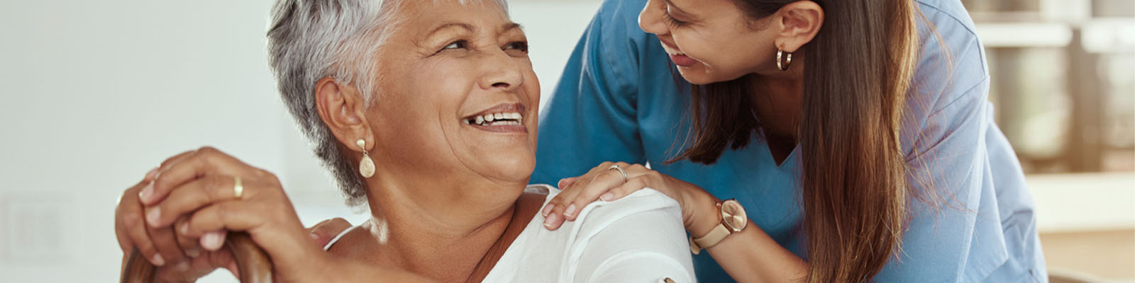 A female senior living nursing assistant with her hands on a female residents' shoulders as they smile at each other