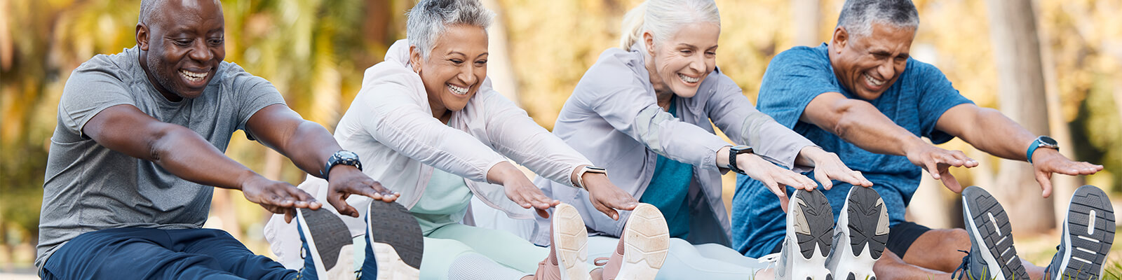 Two male and two female senior living residents dressed in athletic clothes sitting on the ground smiling while reaching their arms out to touch their toes
