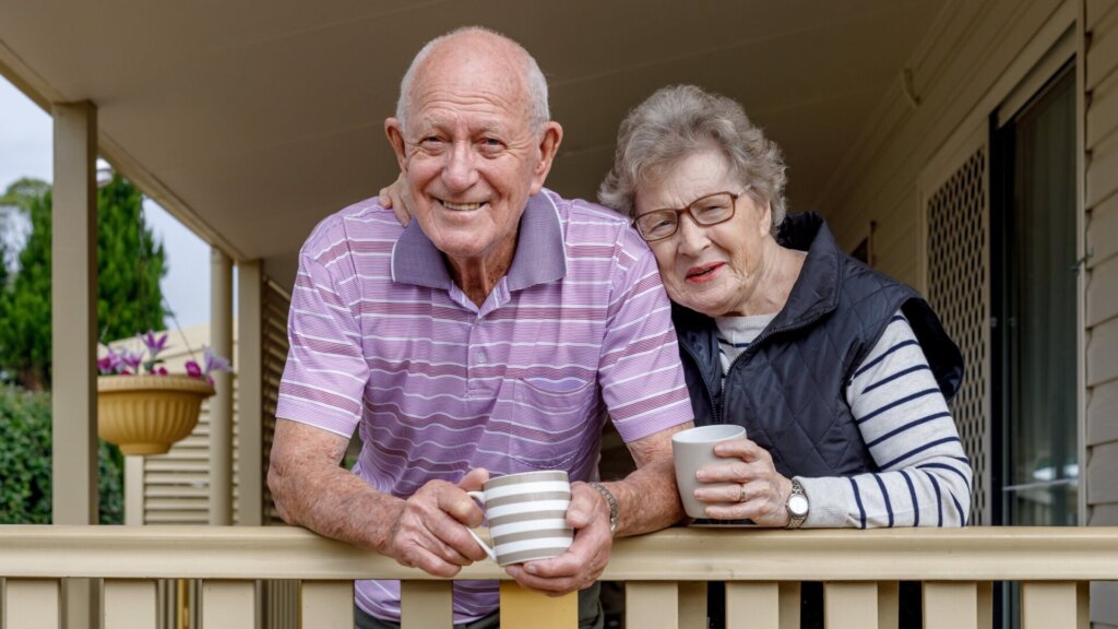 A senior man and senior woman standing together on their front porch while leaning against the railing and holding mugs in their hands