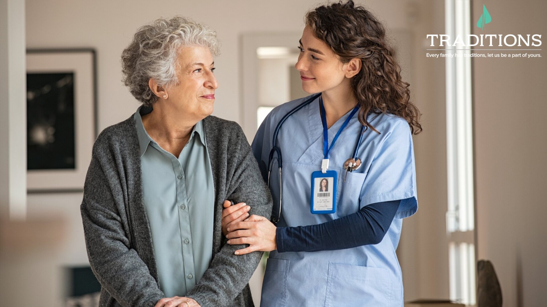 A senior woman standing and looking over at her female nurse who is stabilizing her to help her stand up with the Traditions logo in the top right corner. Quincy Place Senior Living.