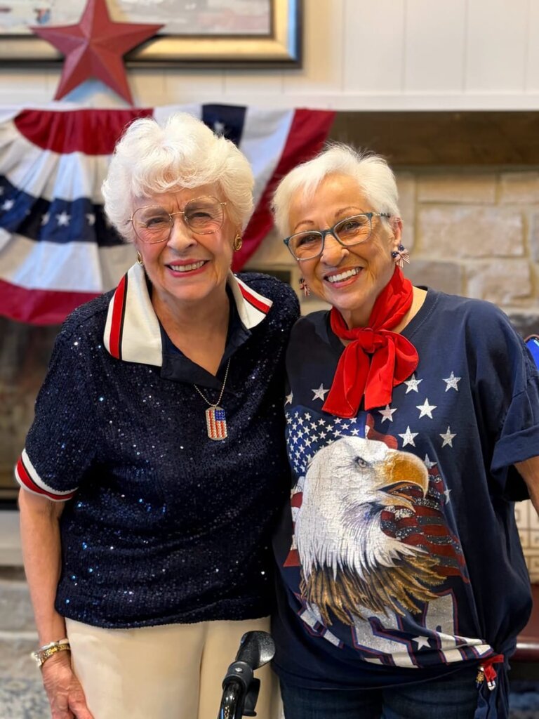 Two residents in patriotic outfits posing together in front of a fireplace decorated with a flag bunting for the Fourth of July